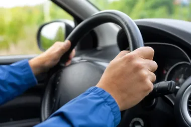 Kierowca w niebieskiej kurtce trzyma dłoniom czarną kierownicę samochodu osobowego.