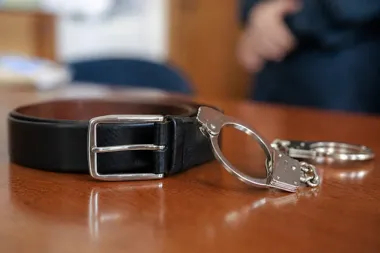 Kajdanki i czarny pasek leżą na drewnianym stole, w tle rozmyta postać siedzącej osoby.
